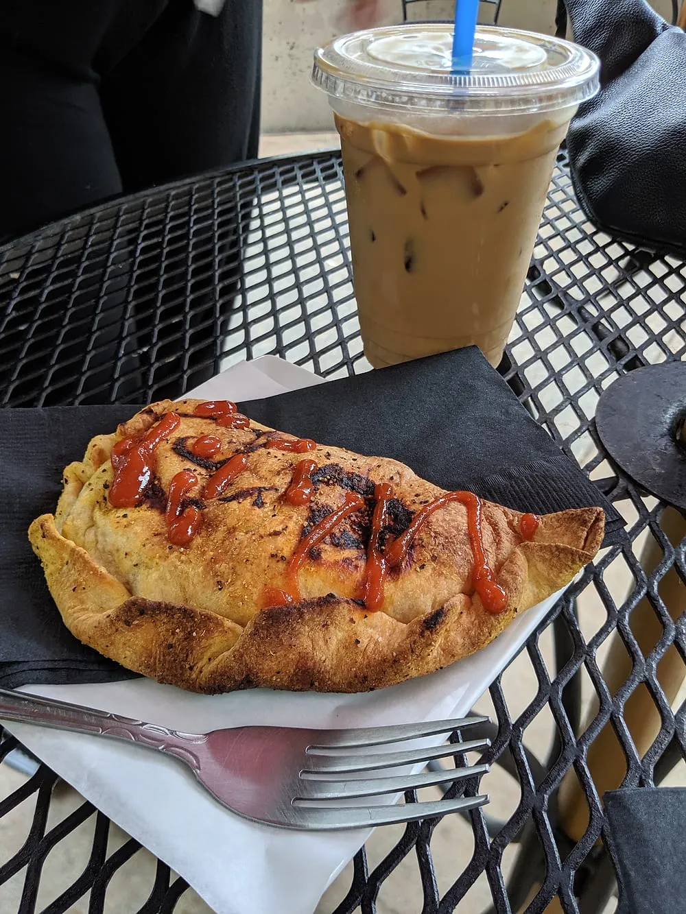 A samosa and an iced coffee on the black mesh table of the sidewalk patio.
