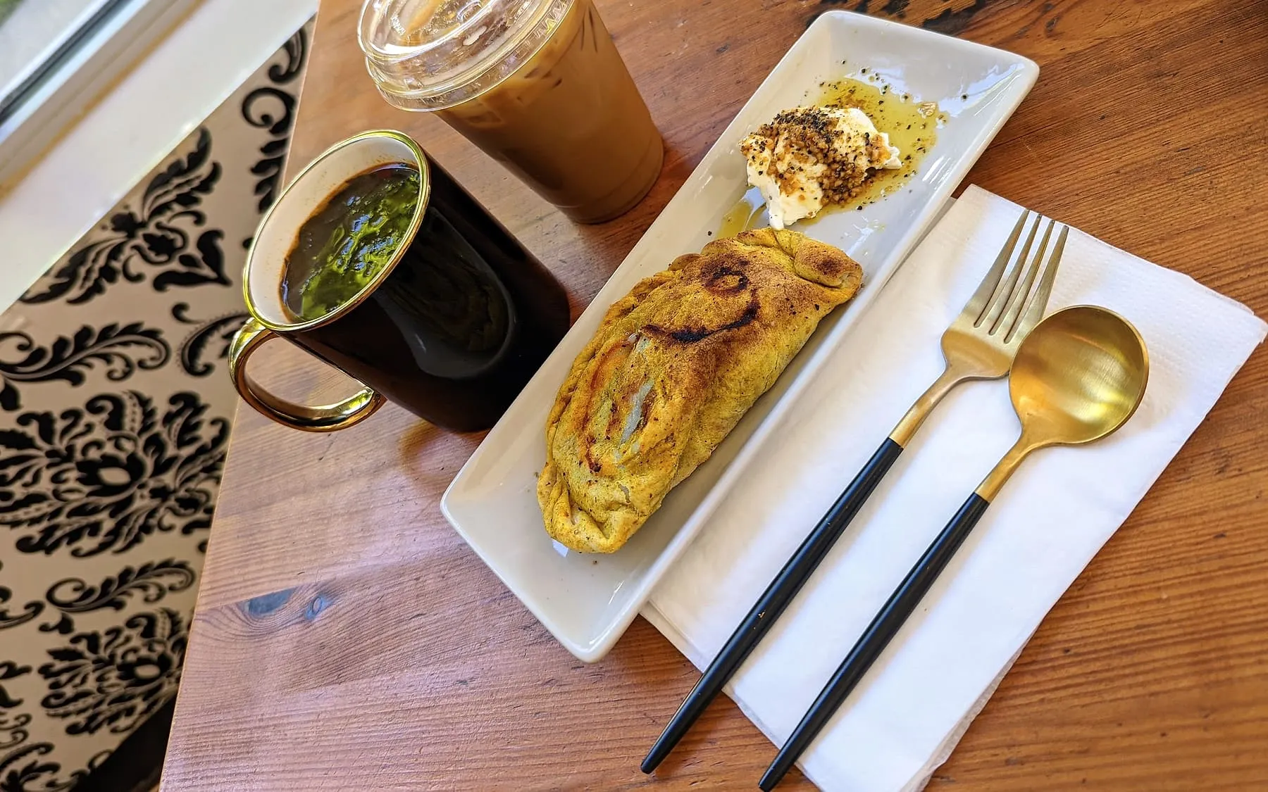 An iced latte, a mug of black coffee and a samosa on a wooden table beside a damask runner at Cafe Argento.